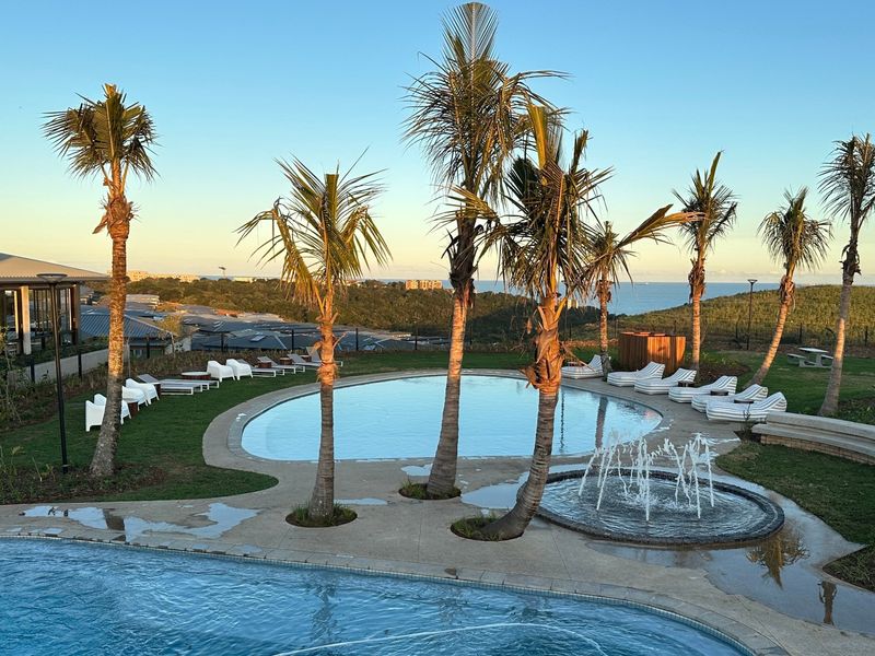 A well-maintained outdoor pool area featuring a swimming pool, water fountain, landscaped garden, and paved deck with scenic coastal views.