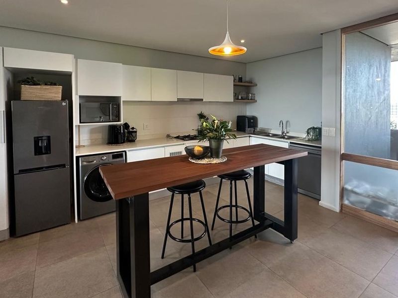 Modern kitchen featuring sleek white cabinetry, integrated appliances, a built-in hob, and a central breakfast bar, seamlessly connected to an outdoor balcony area.