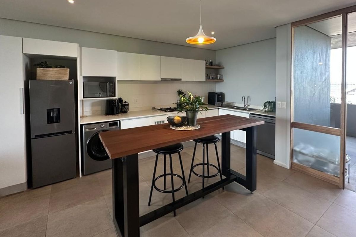 Modern kitchen featuring sleek white cabinetry, integrated appliances, a built-in hob, and a central breakfast bar, seamlessly connected to an outdoor balcony area.
