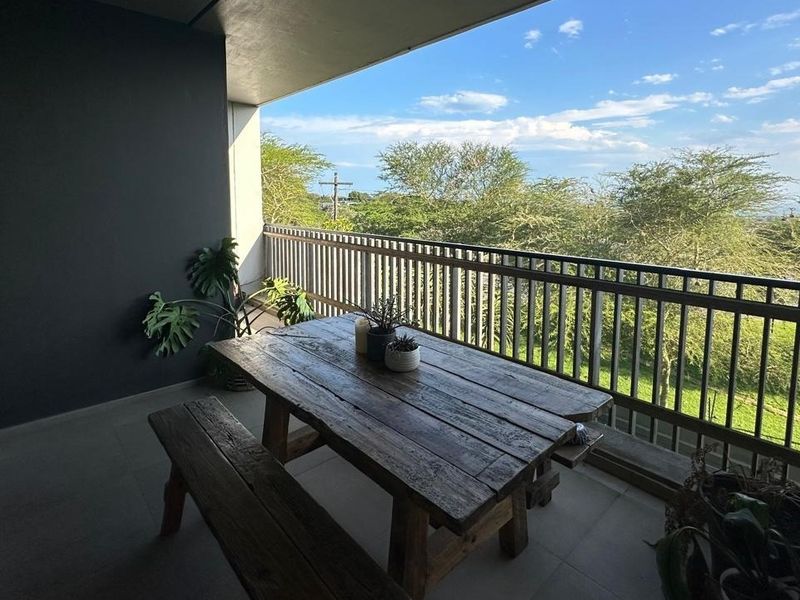 A spacious covered balcony featuring tiled flooring and a metal balustrade, offering pleasant views of the surrounding greenery.