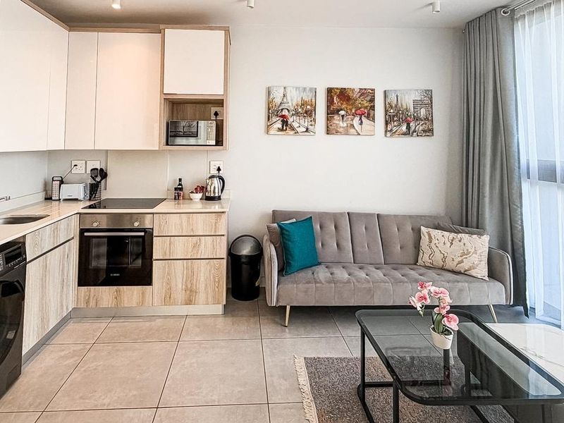Modern open-plan apartment featuring a compact kitchen with built-in appliances, tiled flooring, and a bright living area with floor-to-ceiling window access.