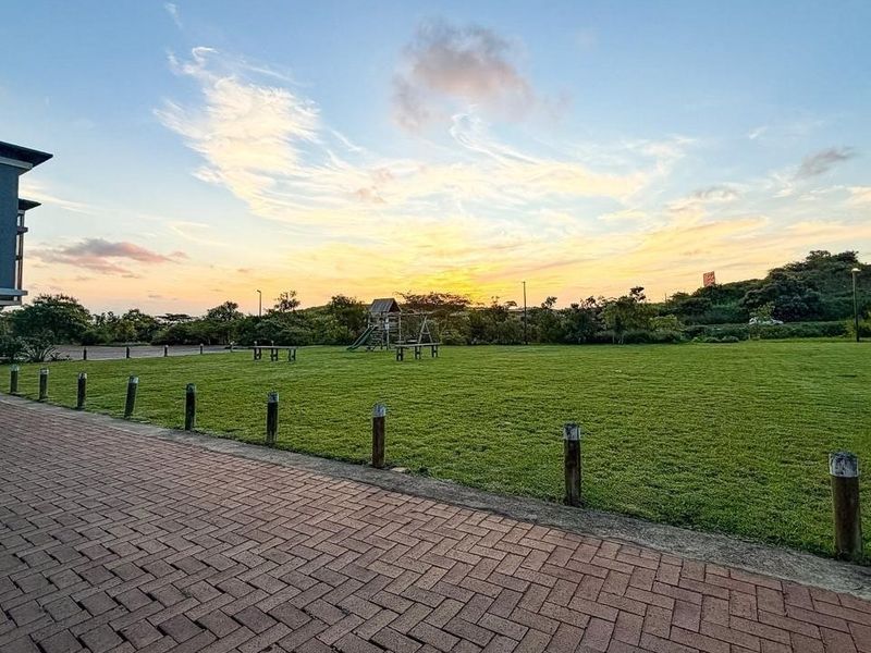 A well-maintained communal garden area featuring a paved walkway, timber bollards, and expansive lawn space at sunset.