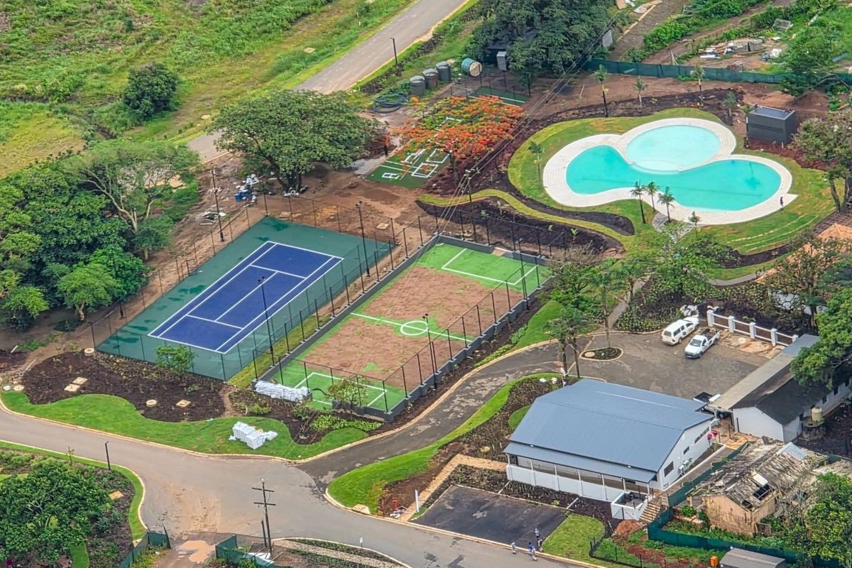 Aerial view of the property grounds featuring a tennis court, multi-purpose sports court, swimming pool, and paved access areas within a landscaped estate.