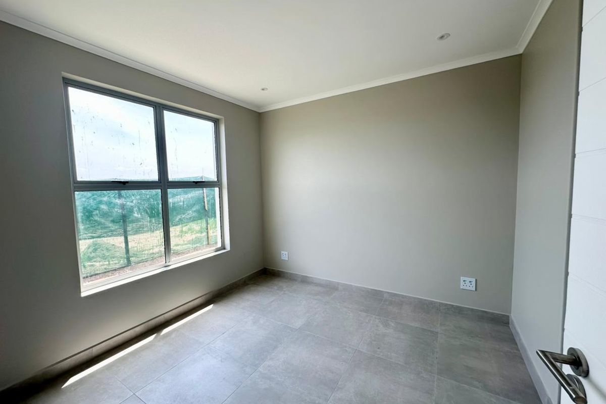 A bright, empty bedroom featuring neutral wall tones, large aluminium-framed windows, and modern grey floor tiles.