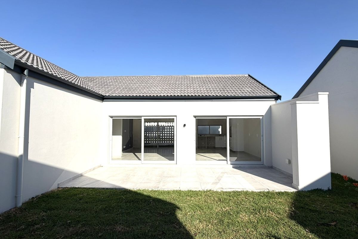 A well-maintained rear exterior view featuring a paved patio area, sliding glass doors, and a neat lawn under a clear blue sky.