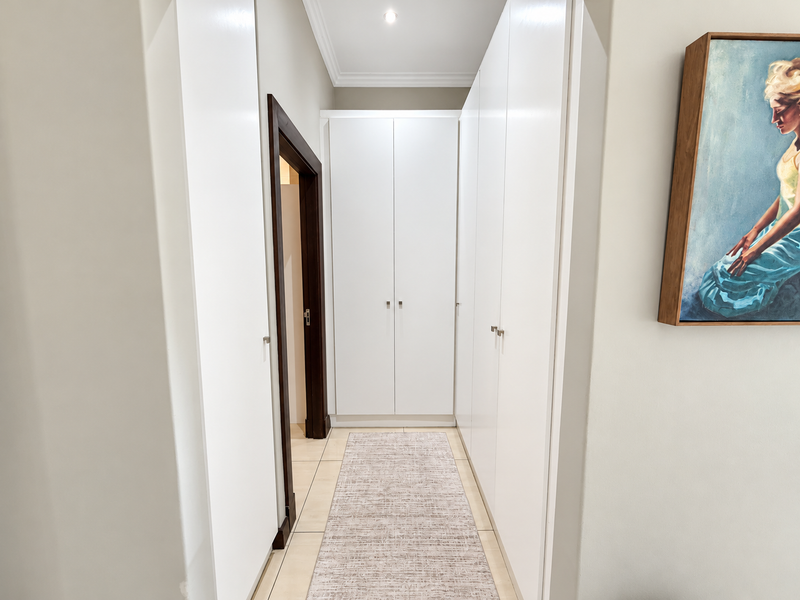 A clean, well-maintained hallway featuring extensive built-in storage cupboards, neutral tiled flooring, and modern recessed lighting.