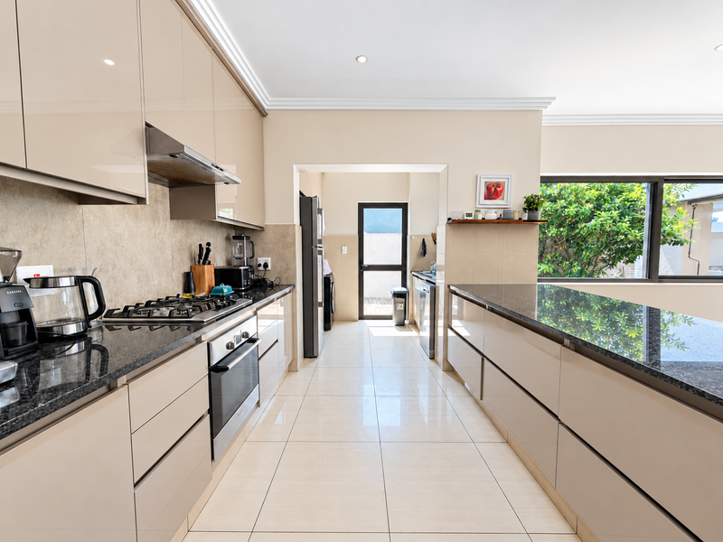 Modern kitchen featuring high-gloss cabinetry, granite countertops, integrated appliances, and tiled flooring, leading to a scullery area with exterior access.