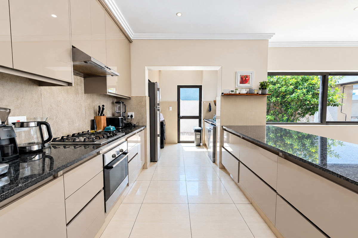 Modern kitchen featuring high-gloss cabinetry, granite countertops, integrated appliances, and tiled flooring, leading to a scullery area with exterior access.