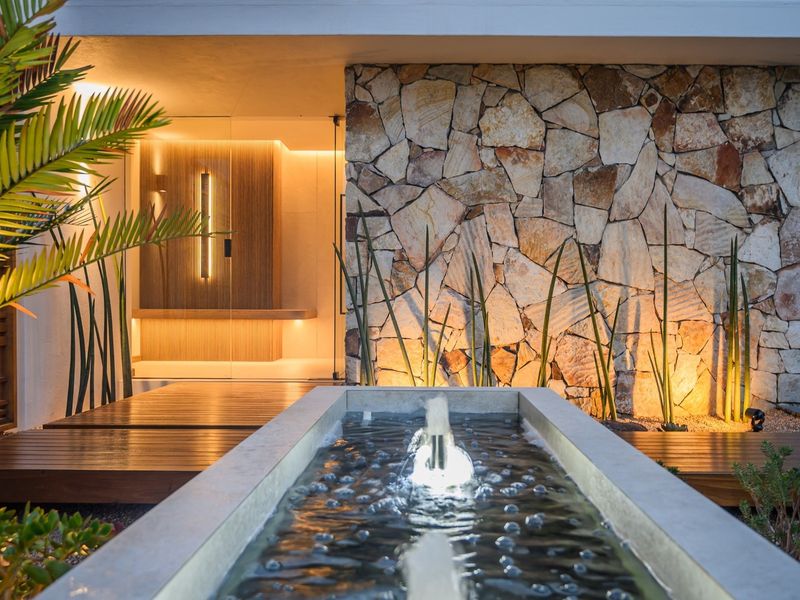 A sophisticated entrance featuring a modern water feature, natural stone cladding, timber decking, and warm architectural lighting.