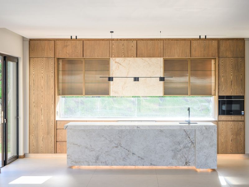 Modern kitchen featuring a large marble island, custom wood cabinetry, integrated appliances, and elegant under-cabinet lighting with a clean, minimalist aesthetic.