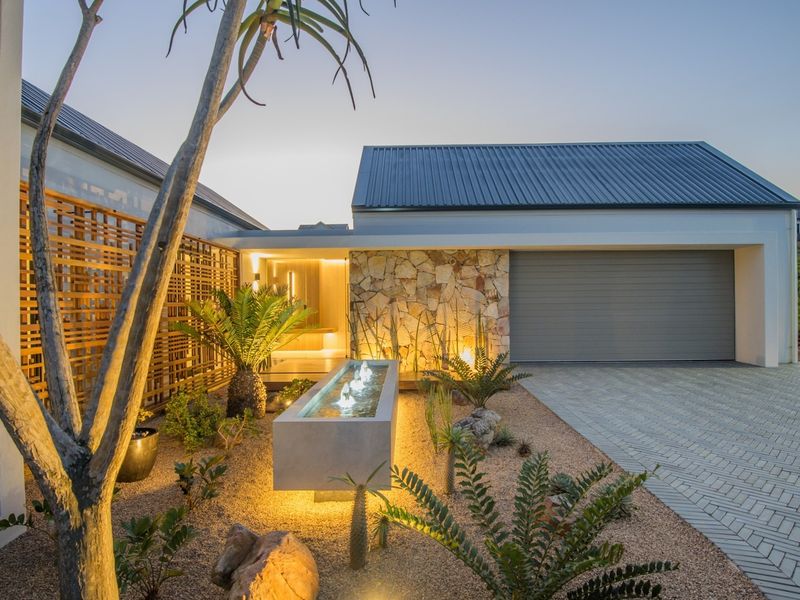 Modern architectural front facade featuring stone cladding, a sleek garage door, a landscaped garden with a water feature, and ambient exterior lighting.