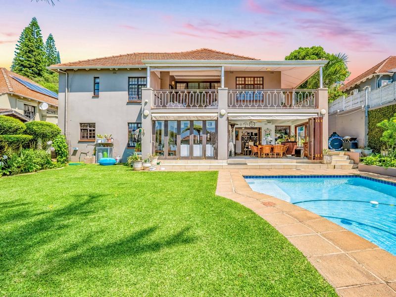 Rear view of a two-story house featuring a swimming pool, landscaped garden, covered patio, and an upper-level balcony with wrought iron railings.