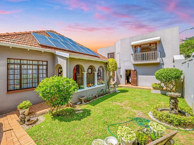 Rear view of the property featuring a landscaped garden, paved walkways, an arched veranda, and a balcony, with solar panels installed on the roof.
