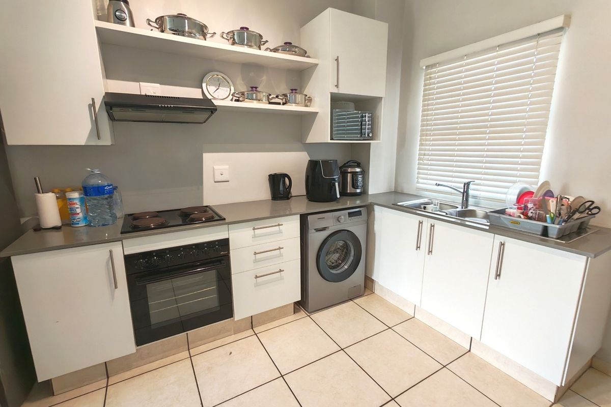 A functional kitchen featuring white cabinetry, dark countertops, an integrated washing machine, electric hob, oven, and a double sink with venetian blinds.