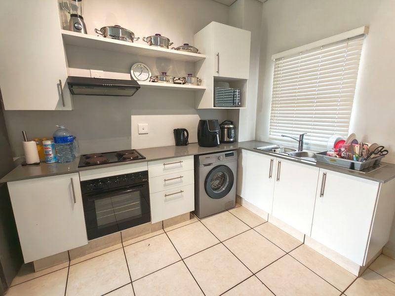 A functional kitchen featuring white cabinetry, dark countertops, an integrated washing machine, electric hob, oven, and a double sink with venetian blinds.