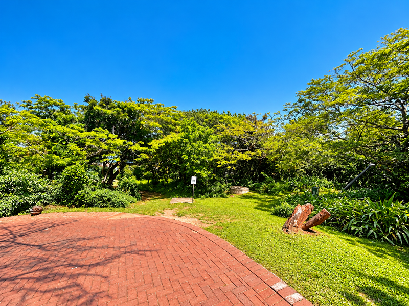 A well-maintained residential garden featuring a brick-paved patio area, lush green lawn, and mature trees providing natural shade.