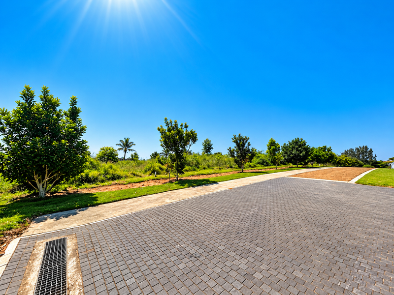 A well-maintained paved driveway leading into a vacant residential plot with established trees and clear blue skies.