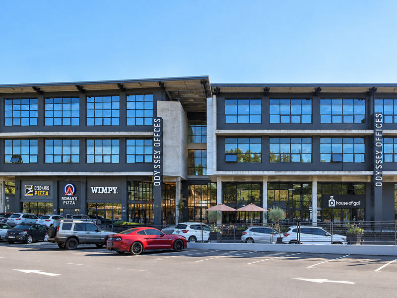 Modern multi-story commercial building featuring a contemporary concrete and glass facade with ground-floor retail spaces and an adjacent paved parking lot.