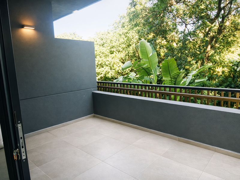 A modern, clean balcony featuring tiled flooring, a sleek metal balustrade, and a wall-mounted light, overlooking lush greenery.