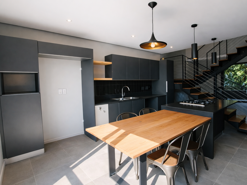 Modern open-plan kitchen featuring dark cabinetry, a central island with a gas hob, and a sleek staircase, finished with contemporary tiled flooring.