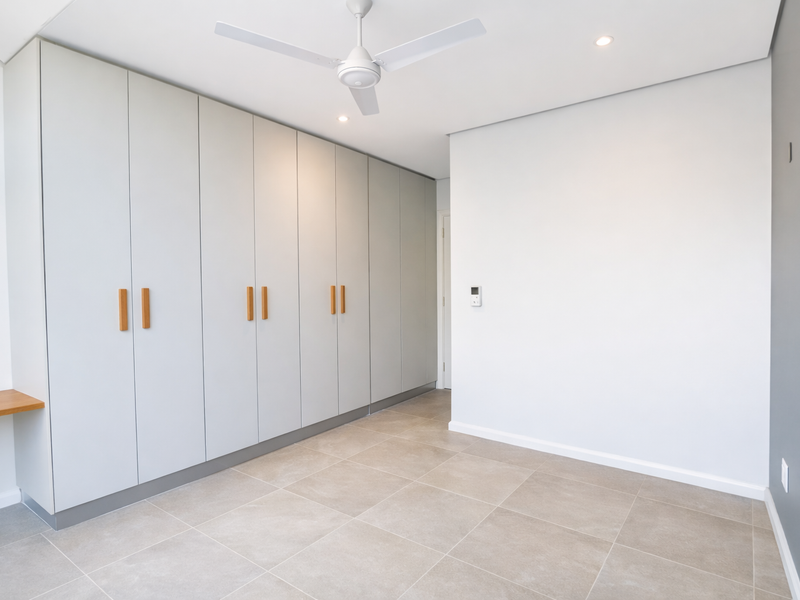 A bright, modern bedroom featuring extensive built-in cabinetry, neutral tiled flooring, a ceiling fan, and recessed lighting in a clean, minimalist design.