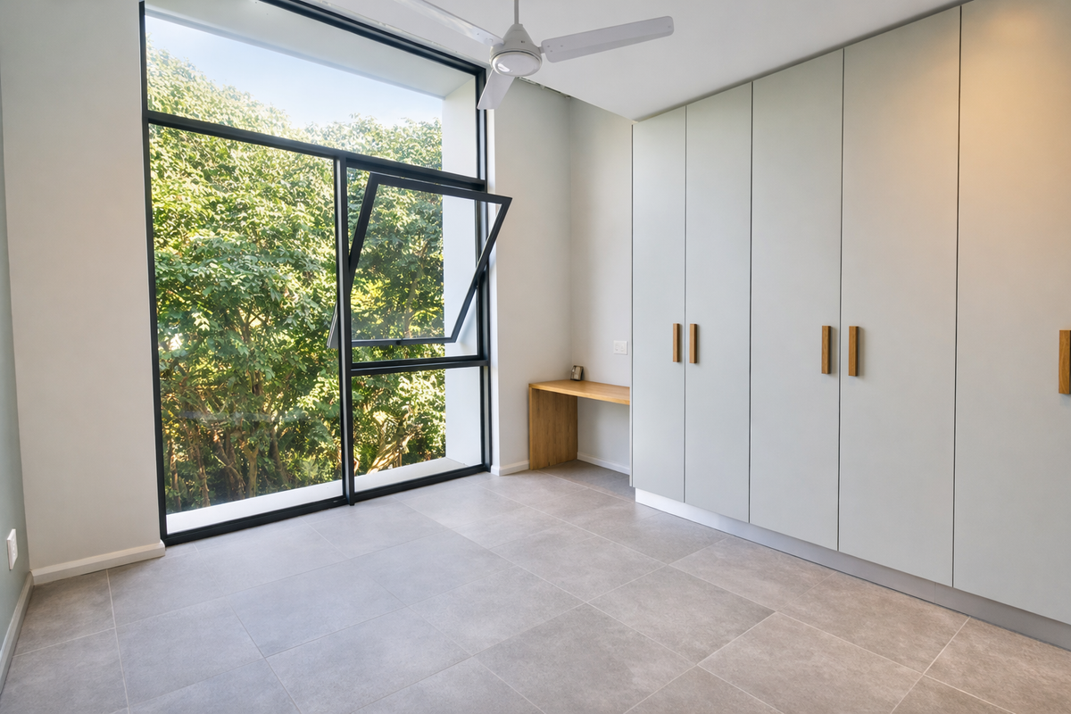 A bright, modern bedroom featuring large windows with leafy views, built-in cupboards, a integrated desk, and neutral tiled flooring.