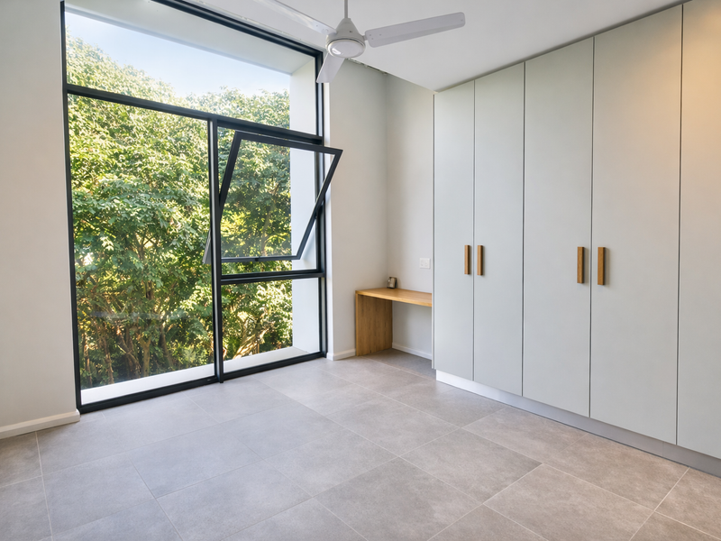 A bright, modern bedroom featuring large windows with leafy views, built-in cupboards, a integrated desk, and neutral tiled flooring.