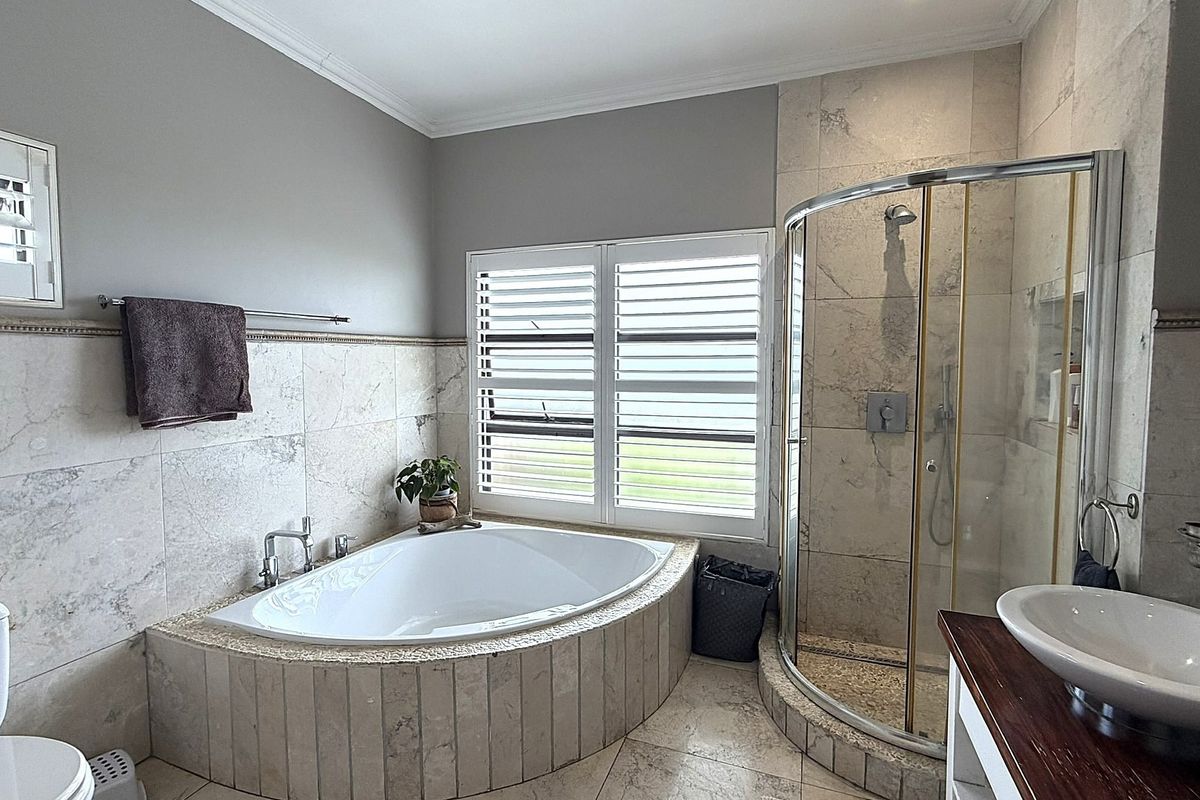 A well-appointed bathroom featuring a corner bathtub, a curved glass shower enclosure, stone tiling throughout, and plantation shutters for natural light control.