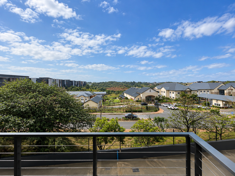 A scenic elevated view overlooking a residential estate with landscaped grounds, paved roads, and surrounding greenery, captured from a balcony with a metal balustrade.