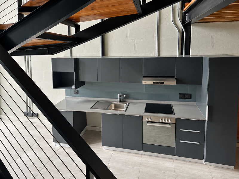Modern industrial-style kitchen featuring dark matte cabinetry, integrated stainless steel appliances, glass splashback, and tiled flooring, situated beneath a feature staircase.