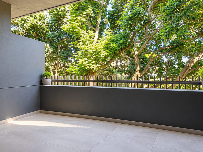 A modern, clean balcony featuring neutral floor tiles, a sleek metal balustrade, and a wall-mounted light fixture, overlooking lush green trees.