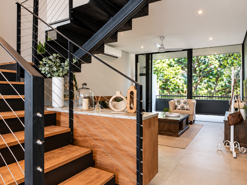 Modern open-plan living area featuring a striking timber staircase with steel cable balustrades, tiled flooring, and seamless access to an outdoor balcony.
