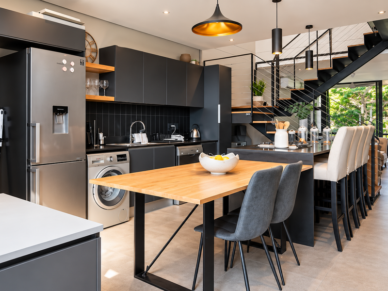 Modern open-plan kitchen and dining area featuring sleek dark cabinetry, integrated appliances, a central island, and a striking staircase leading to the upper level.