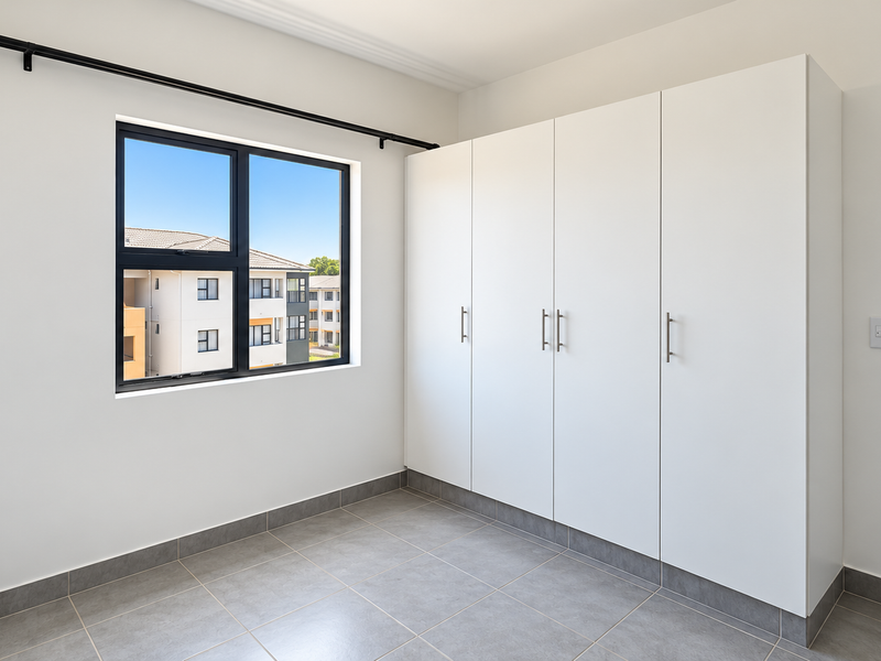 A bright, empty bedroom featuring modern tiled flooring, built-in white cupboards, and an aluminium-framed window with a curtain rod.