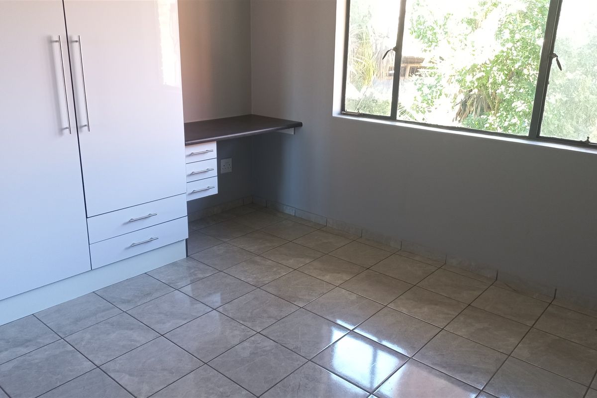 A clean, empty bedroom featuring tiled flooring, a built-in wardrobe, and a fitted desk area with natural light from the window.