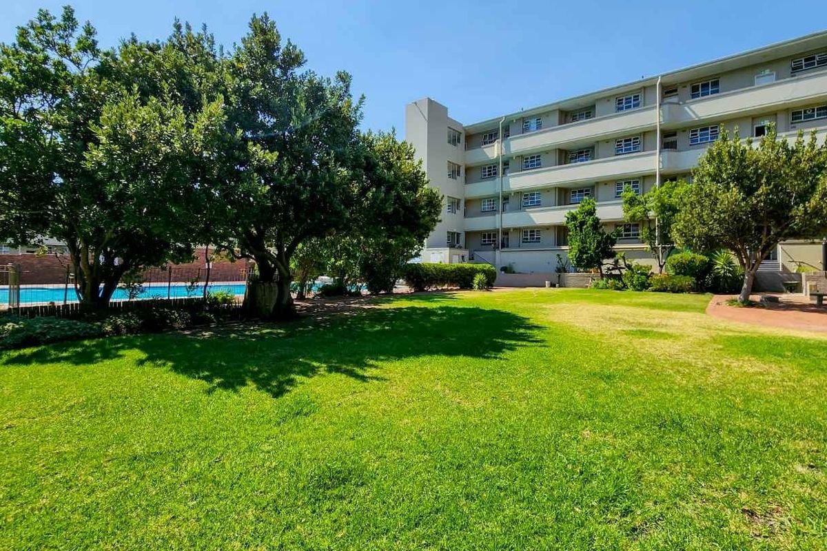 A well-maintained communal garden area featuring a swimming pool, mature trees, and paved patio space set against the apartment building facade.