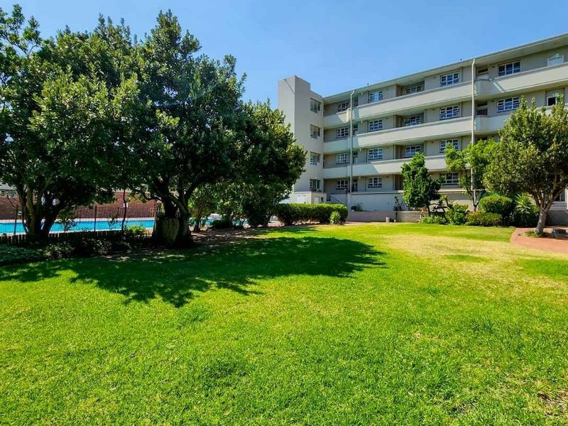 A well-maintained communal garden area featuring a swimming pool, mature trees, and paved patio space set against the apartment building facade.