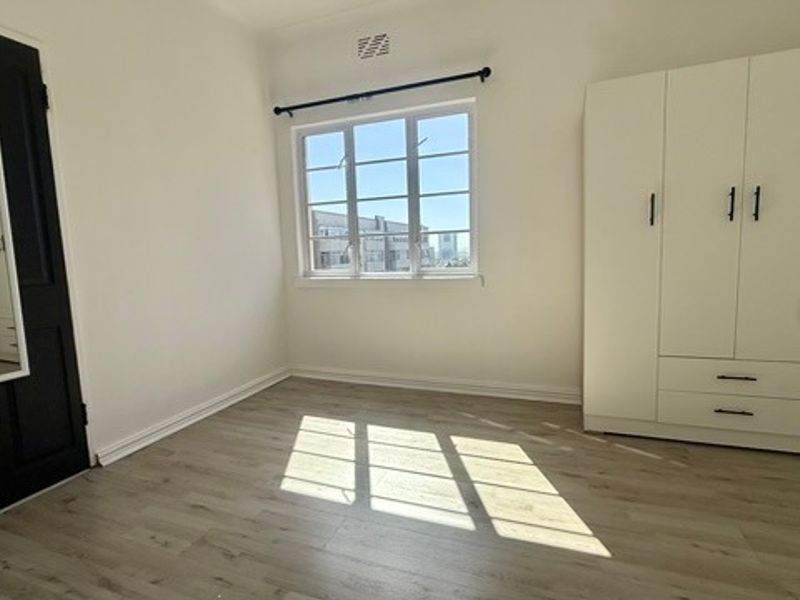 A bright, empty bedroom featuring modern laminate flooring, a large built-in wardrobe, and a window providing natural light.