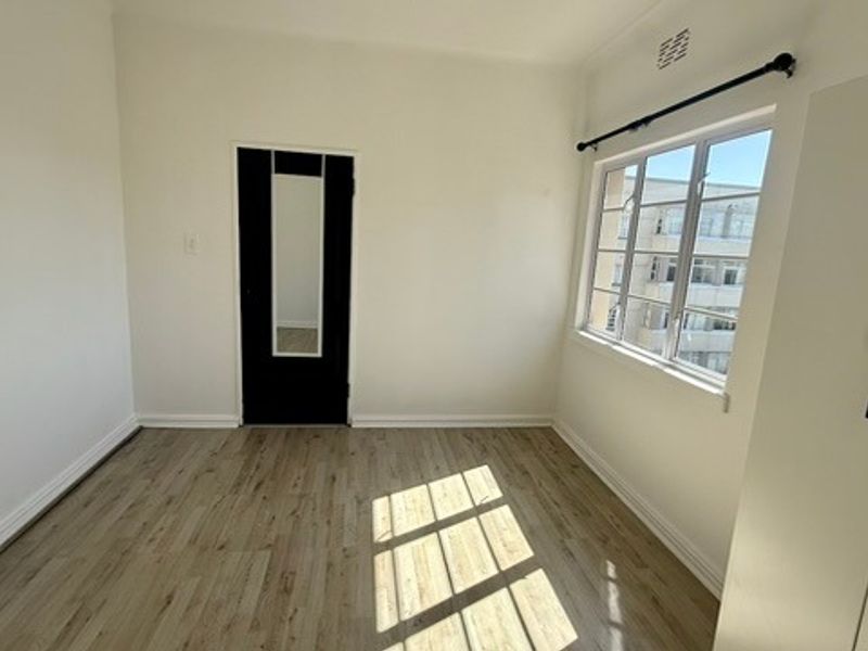 A bright, empty bedroom featuring light-toned laminate flooring, a built-in wardrobe, and a window with security bars.