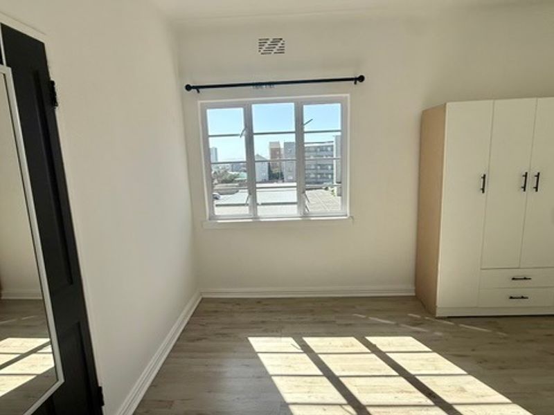 A bright, empty bedroom featuring light laminate flooring, a built-in wardrobe, and a window with views of the surrounding urban area.