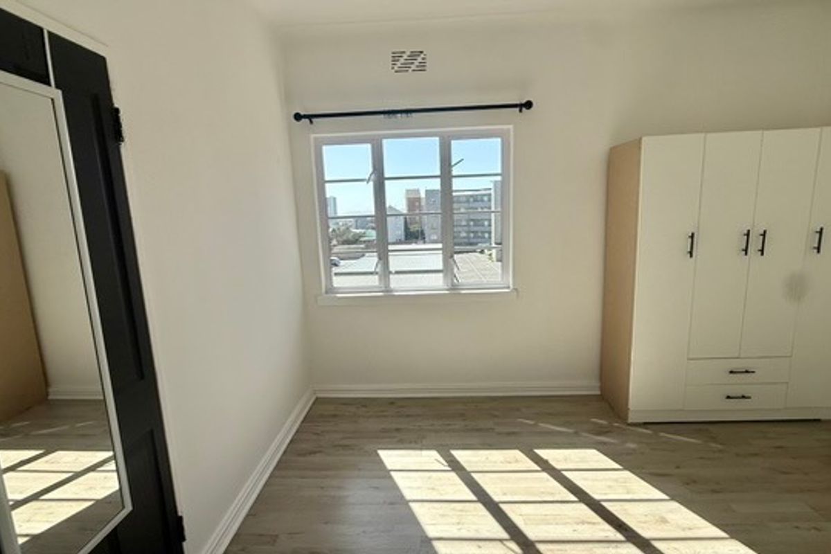 A bright, empty bedroom featuring light laminate flooring, a built-in wardrobe, and a window with views of the surrounding urban area.