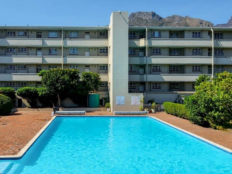 A well-maintained communal swimming pool area set within a multi-story apartment complex, featuring paved surrounds and established garden landscaping.