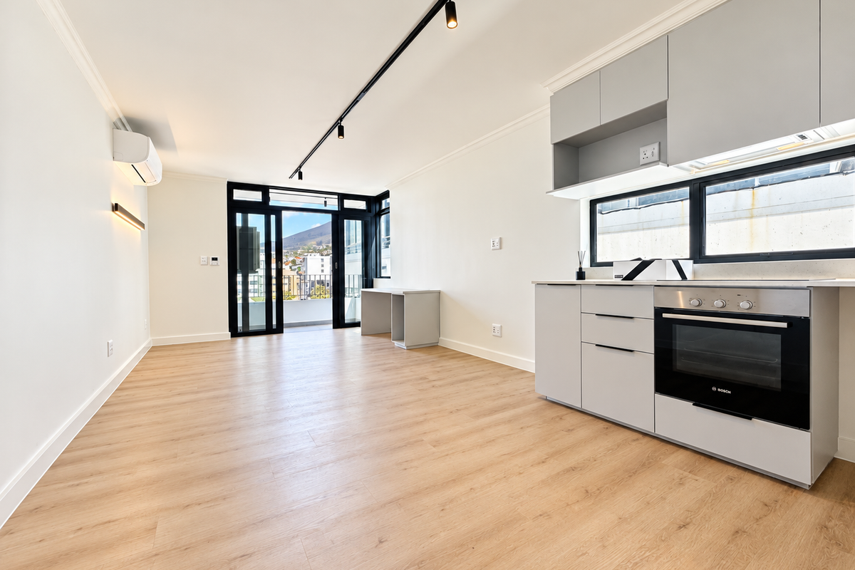 Modern open-plan living area featuring light wood-look flooring, sleek grey cabinetry with integrated oven, track lighting, and direct access to a balcony.