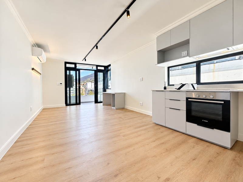 Modern open-plan living area featuring light wood-look flooring, sleek grey cabinetry with integrated oven, track lighting, and direct access to a balcony.