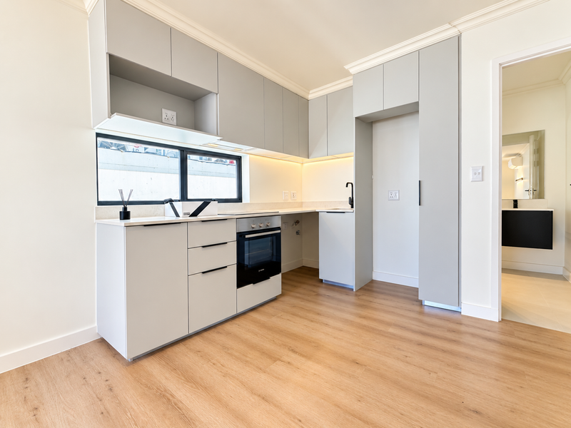 A modern, minimalist kitchen featuring light grey cabinetry, integrated oven, stone countertops, and warm under-cabinet lighting, set against light wood-look flooring.