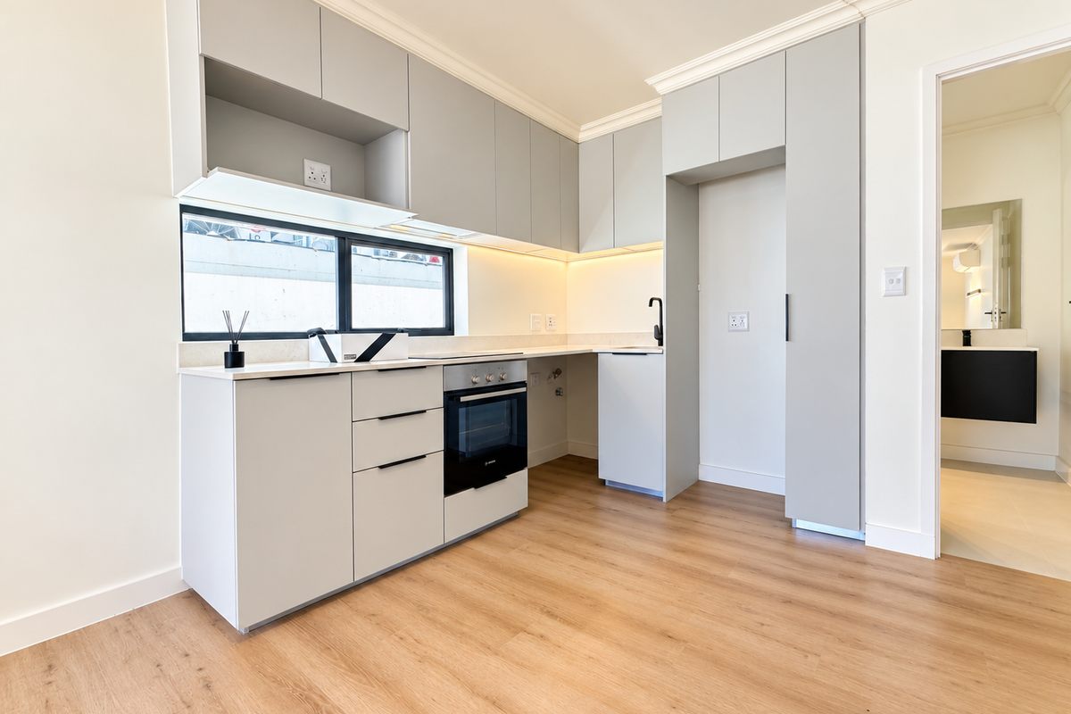 A modern, minimalist kitchen featuring light grey cabinetry, integrated oven, stone countertops, and warm under-cabinet lighting, set against light wood-look flooring.