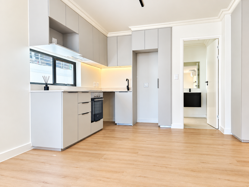 Modern kitchen featuring light grey cabinetry, integrated oven, stone countertops, and warm wood-look flooring, with an adjacent doorway leading to a bathroom.