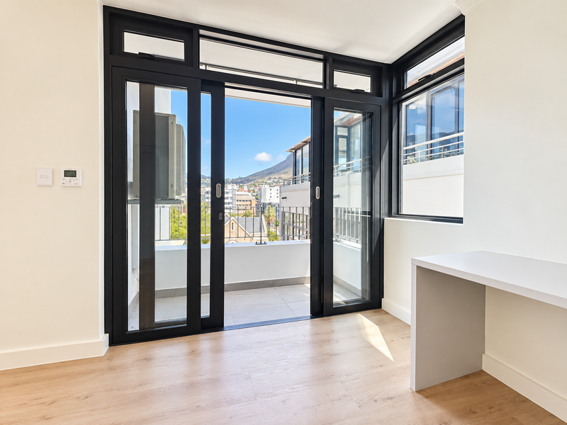 A bright, modern living space featuring sleek black-framed sliding doors leading to a balcony, light laminate flooring, and a built-in desk area.