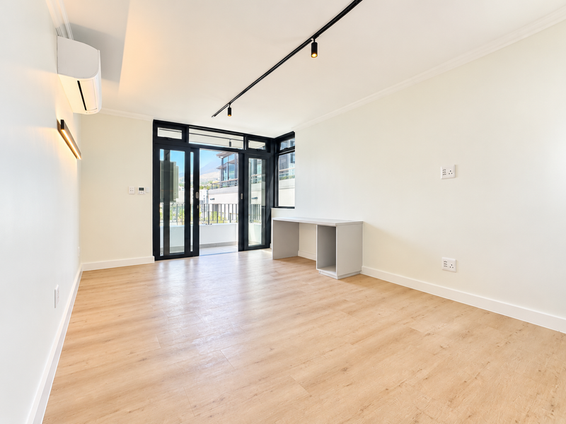 A bright, modern living area featuring light laminate flooring, track lighting, a built-in desk, and sliding glass doors leading to a balcony.