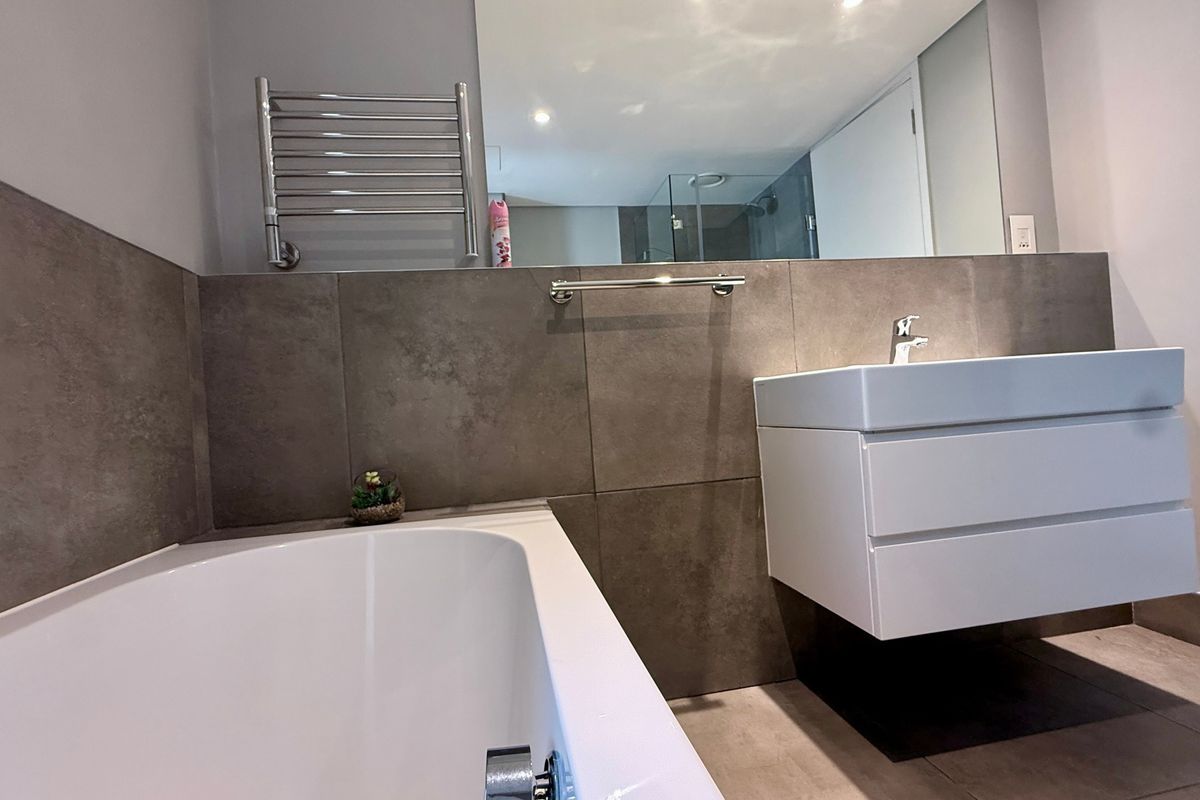 Modern bathroom featuring a wall-mounted vanity, built-in bathtub, heated towel rail, and large wall mirror with neutral grey tiling.